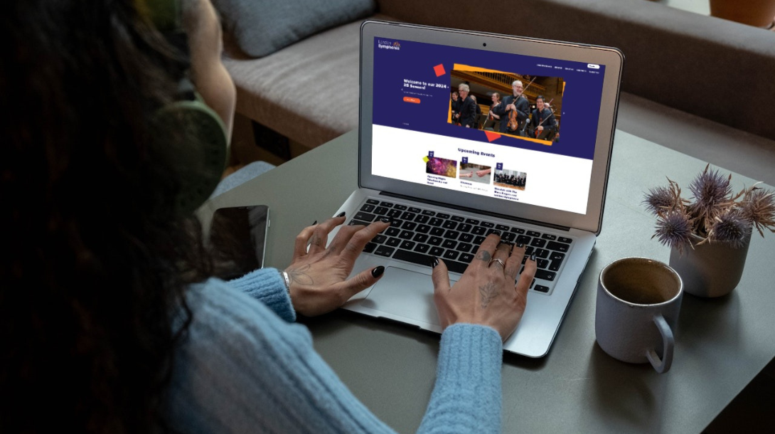 A woman listening to music looking at the London Symphonia website home page on her laptop with a drink and flowers beside her.